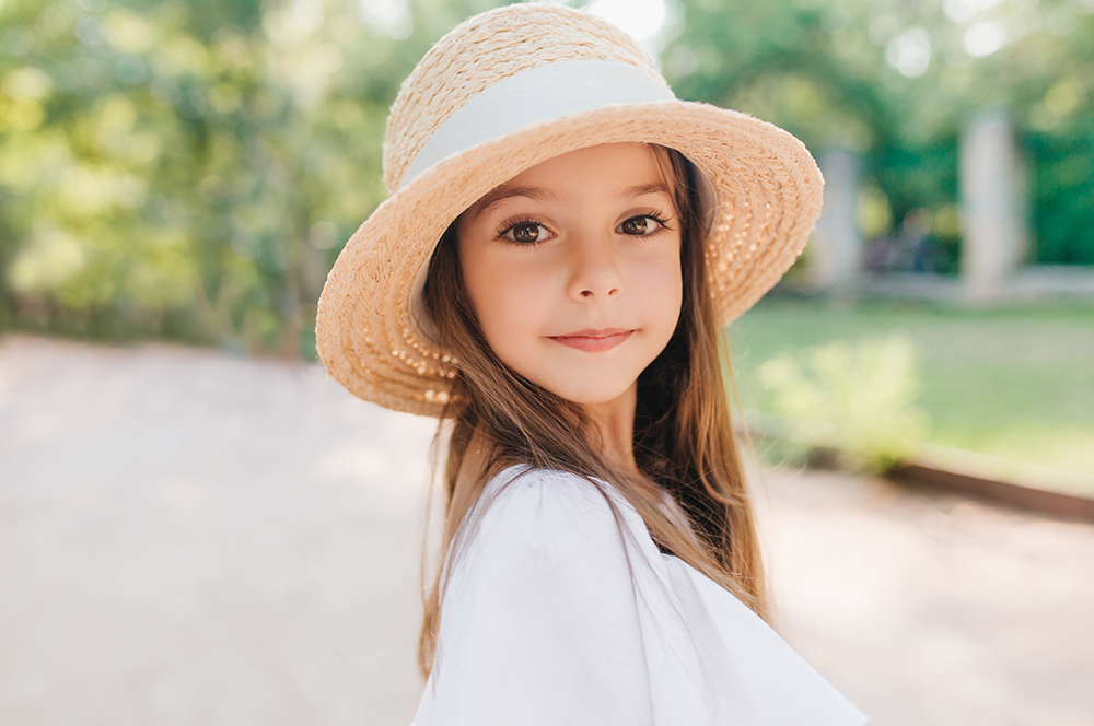Close Up Portrait Of Wonderful Child With Shiny Brown Eyes Looking To Camera With Interest. Enthusiastic Little Girl In Vintage Straw Hat Decorated With Ribbon Posing During Game In Park..