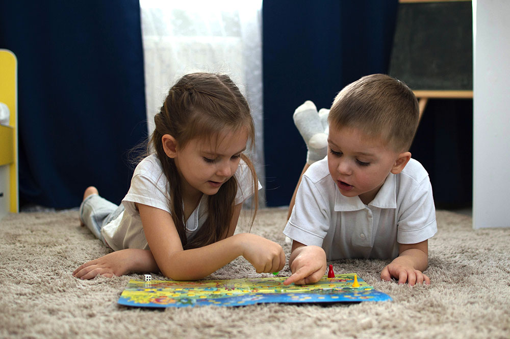 Portrait Of Two Children Lying On The Floor And Playing The Board Game Walker In The Nursery. A Board Game And The Concept Of Children's Leisure. Selective Focus